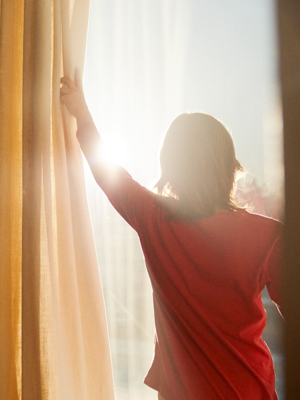 A child in a red t-shirt pulls back yellow and white curtains to reveal the sun blazing through the window.