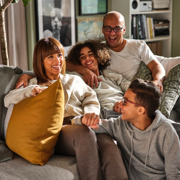 A mum, dad and their two teenage sons, smiling and sitting closely together on a sofa with large cushions in a living room.