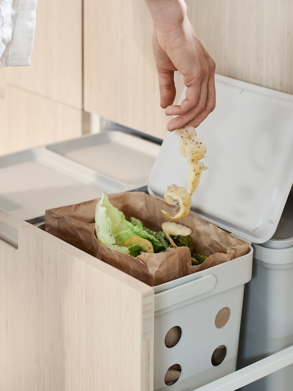 A person adds a vegetable peel to a brown-paper food waste bag in a HÅLLBAR waste sorting bin within a high MAXIMERA drawer.