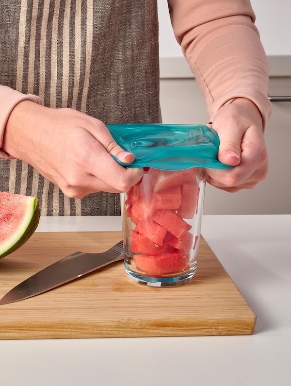 A person places a turquoise, silicone, ÖVERMÄTT food cover on a glass of chopped watermelon that stands on a chopping board.