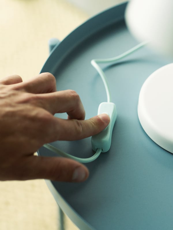 A person’s finger presses the on/off switch on the cord of a white FUBBLA LED work lamp on a light-blue tray table.