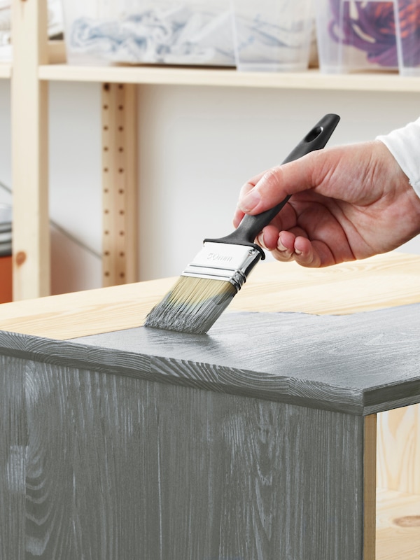 A person’s hand holds a paintbrush and applies grey paint to the top surface of a piece of untreated pine furniture.