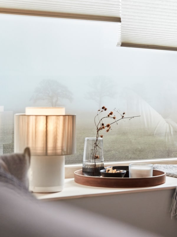 A SYMFONISK speaker lamp with textile shade on a window sill. Outside there is a tree, barely visible through the mist.