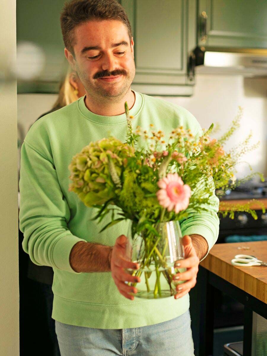A man in a green sweatshirt holds a vase filled with flowers in the kitchen. He is smiling.