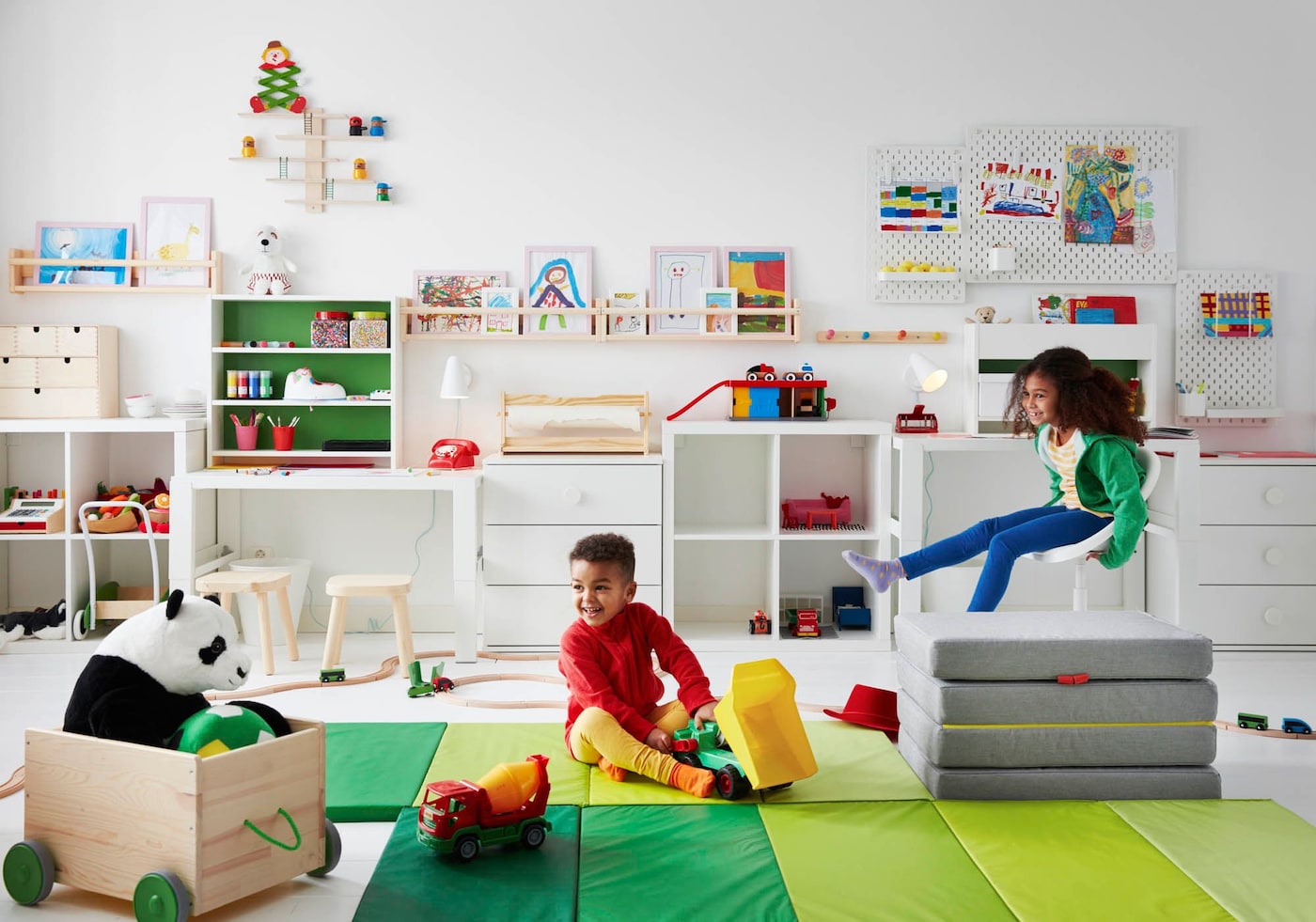 Two children play in a room with green mats on the floor and toys neatly arranged on white shelving on the wall behind.