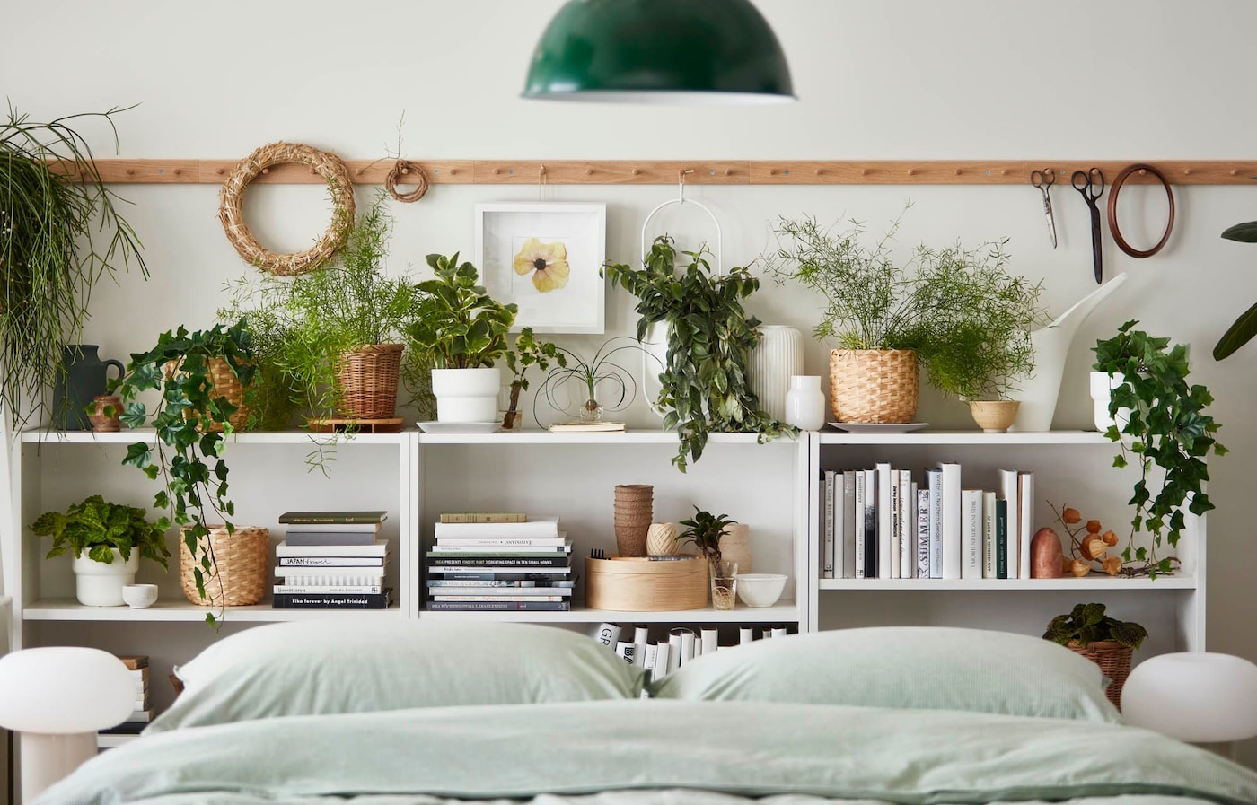 A bedroom with a bed in front of a white shelving unit holding books, plants, boxes and other decorations set against a wall.
