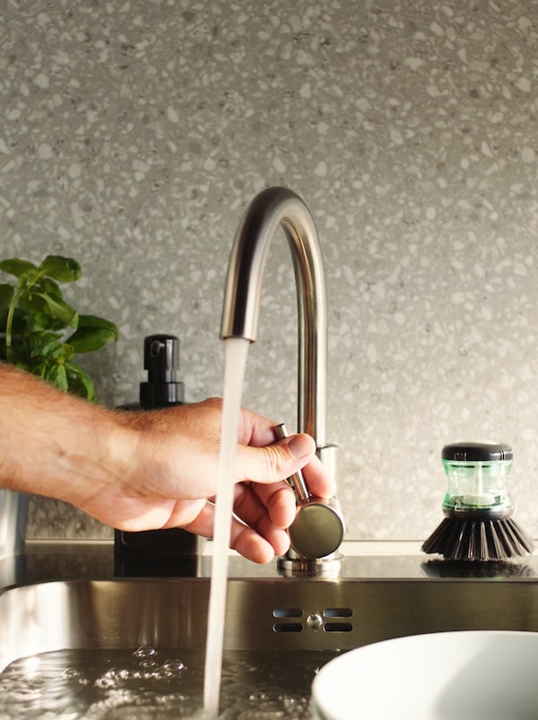 Ruka upravující proud vody kuchyňské baterie GLYPEN z nerezavějící oceli vedle kartáčku na mytí nádobí TÅRTSMET.