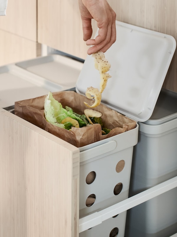 The hand of a person lowering a long string of potato peel into a HÅLLBAR bin containing organic kitchen waste.